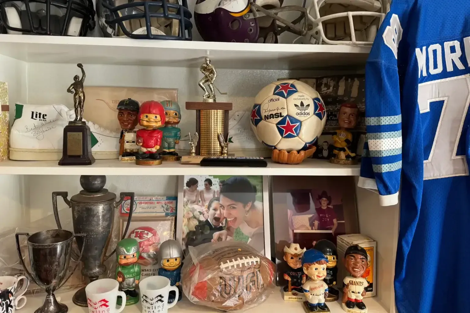Sell game used sports equipment — Shelf of vintage football helmets, an NASL soccer ball, bobbleheads, trophies and silver loving cups