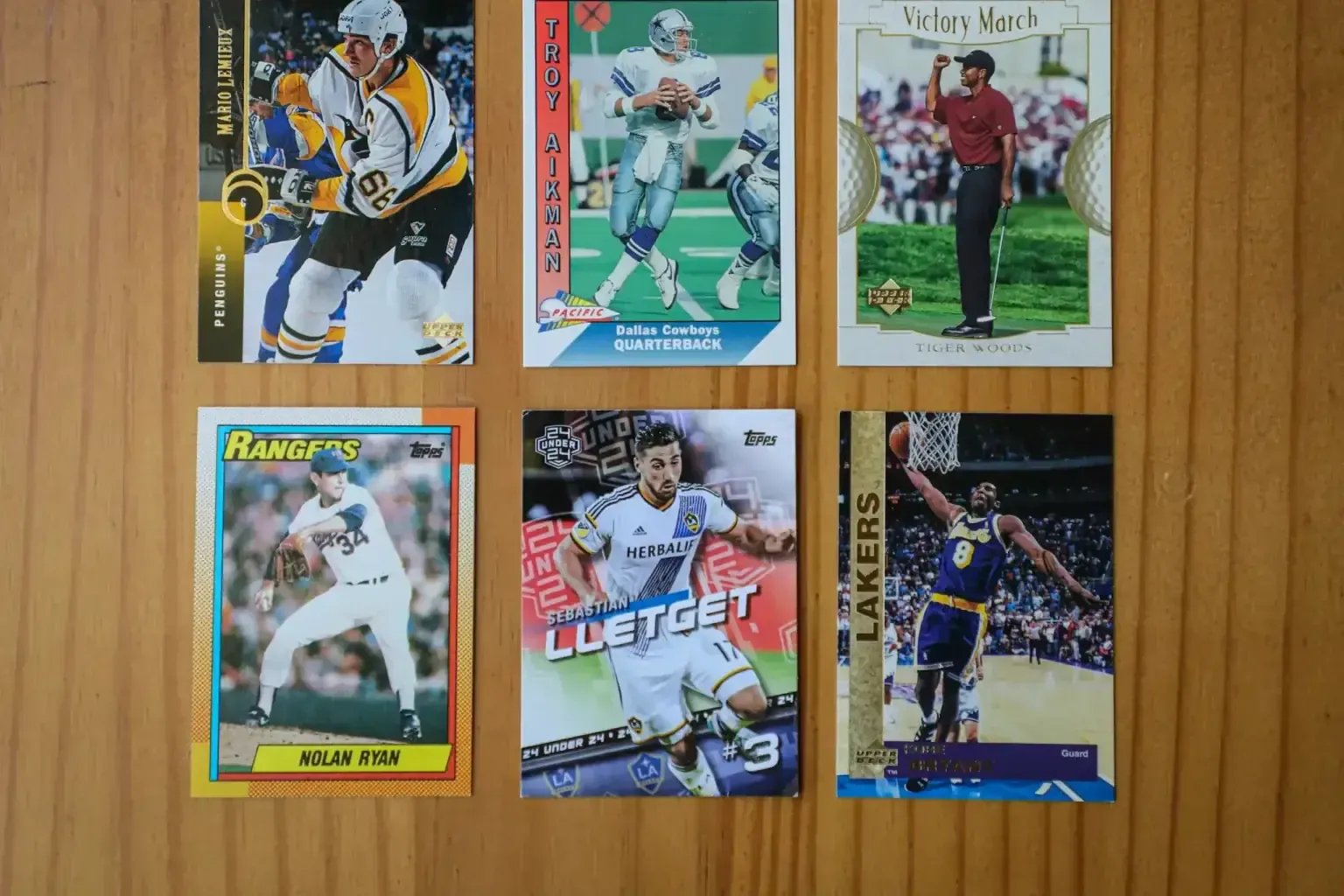 Vintage baseball card collection — Six vintage trading cards laid out on wood: Mario Lemieux hockey, Troy Aikman football, Tiger Woods golf, Nolan Ryan baseball, soccer and Kobe Bryant basketball
