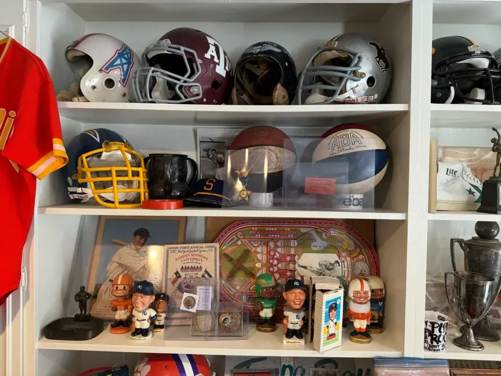 Sell vintage sports memorabilia in San Antonio Tx — Shelf of vintage football helmets, ABA basketballs, bobbleheads and trophies beside a Chiefs jersey